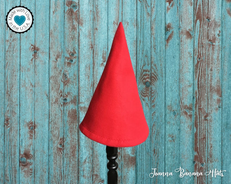 May include: A red felt gnome hat on a wooden stand. The hat is pointy and has a simple design. The stand is black and has a round base.