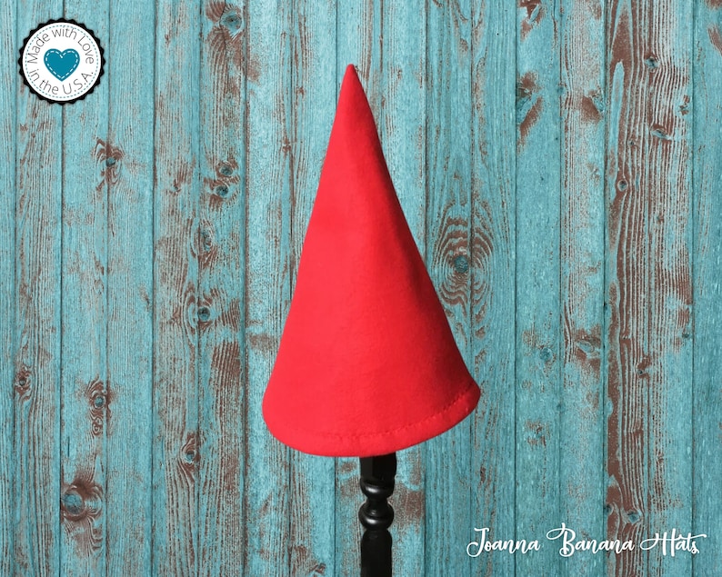 May include: A red felt gnome hat on a wooden stand. The hat is pointy and has a wide brim. The stand is black and has a decorative scroll design.