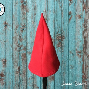 May include: A red felt gnome hat with a pointed top. The hat is sitting on a wooden surface.