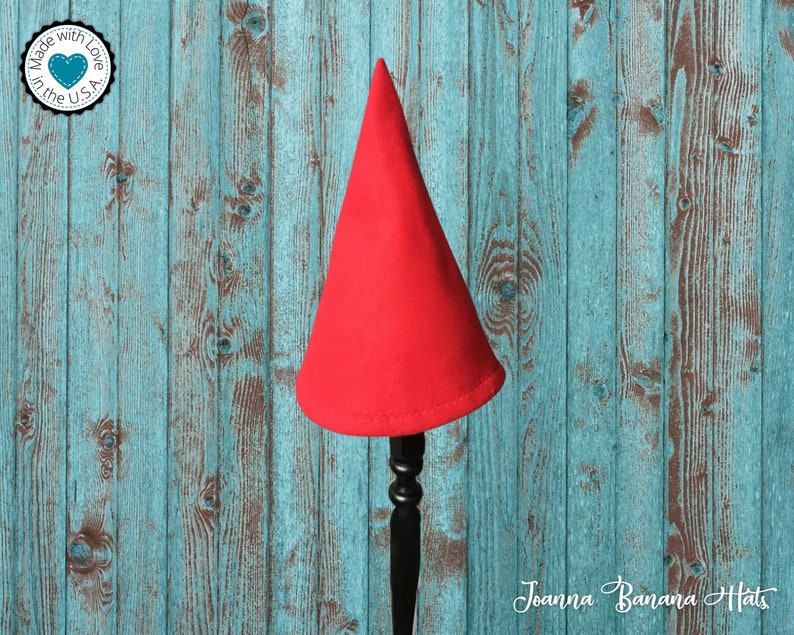 May include: Red felt gnome hat on a wooden stand. The hat is pointy and has a simple design. The stand is black and has a decorative top.