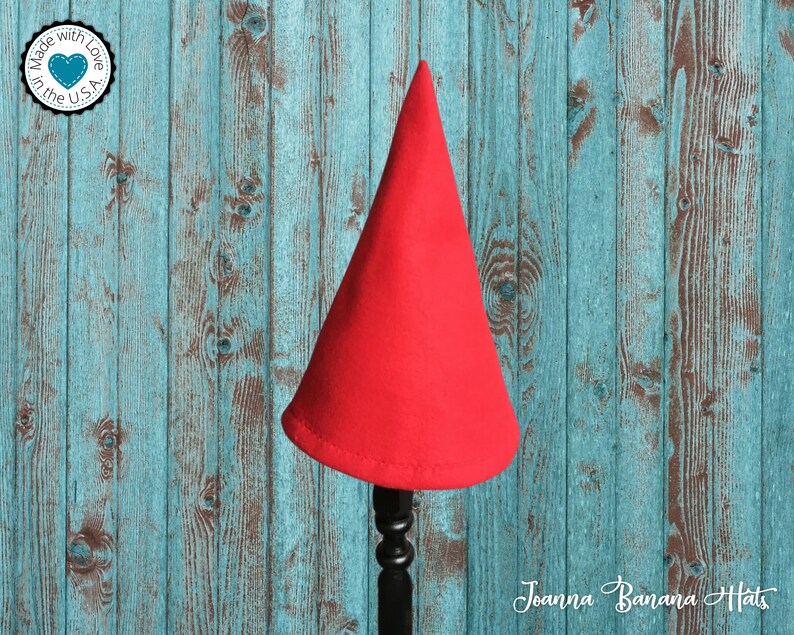 May include: A red felt cone-shaped hat on a wooden stand against a blue and brown woodgrain background. The hat is made in the USA with love.