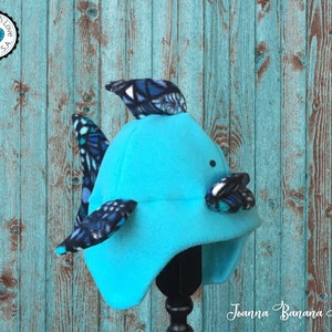 May include: A turquoise blue fleece hat with a fish fin design. The hat has black and blue patterned fabric on the fins. The hat is on a black stand against a turquoise blue wooden background. The text "Joanna Banana Hats" is visible in the bottom right corner of the image.