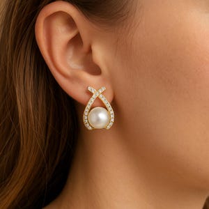 May include: A gold-toned earring with a large, round, white pearl at the center. The earring features a decorative, open design with a crisscross at the top and is encrusted with small, clear crystals. The earring is worn on a person's ear.