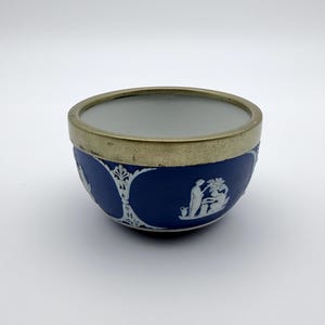May include: A blue ceramic bowl with a gold rim and a light gray interior. The bowl features white relief designs around the exterior, depicting classical figures and decorative elements. The bowl is a decorative item.