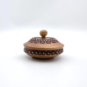 May include: A round wooden lidded container featuring a natural light brown finish. The lid is adorned with a ring of dark red heart-shaped motifs, while the base displays a continuous pattern of dark brown spiral carvings around its circumference.