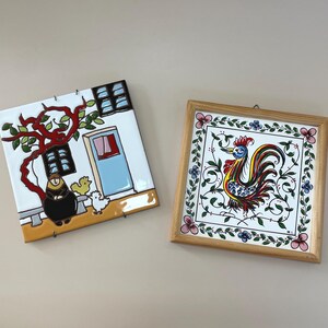 May include: Two square ceramic tiles. One depicts a scene with a building, a tree, and figures in black and yellow. The other tile, framed in wood, features a colorful rooster design with floral accents.