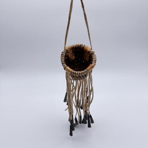 May include: A small, handmade woven basket with a leather strap for hanging. The basket is round with a natural, earthy color palette. Long leather fringe hangs from the bottom, with dark, cone-shaped accents.