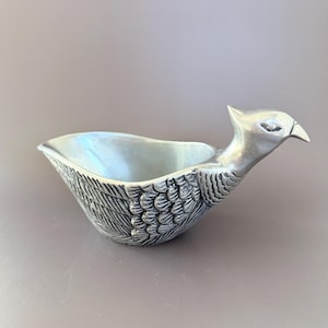 May include: A silver-colored gravy boat in the shape of a bird. The bird's head forms the spout, and the body is the bowl. The bowl has a textured feather design. The gravy boat is made of metal and sits on a neutral background.