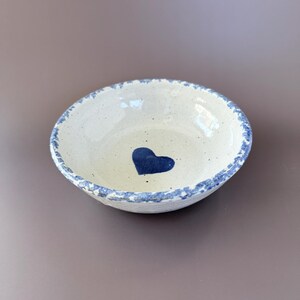May include: A small, off-white ceramic bowl with a blue heart in the center. The rim is decorated with a blue, speckled pattern. The bowl has a rustic, handmade appearance.