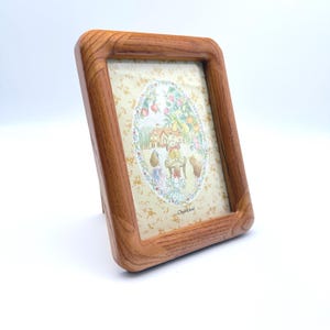May include: A rectangular wooden picture frame with a light brown grain finish. The frame holds a print featuring a yellow background with a small floral pattern and a central oval illustration of a house, trees, and figures with the text Cherished.