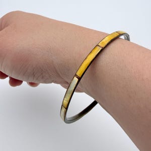 May include: A thin, circular bangle bracelet featuring a dark metal band inlaid with alternating rectangular segments of iridescent white and golden-yellow shell material, creating a smooth, continuous surface around the wrist.