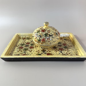May include: A decorative, hand-painted serving set featuring a round lidded bowl and a rectangular tray. The set is painted with a floral design in red, green, and gold on a cream background. The tray has a black base and a cream-colored border.
