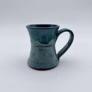 May include: A teal ceramic mug with a curved handle. The mug features a flared top and a textured body with embossed swirl designs. The mug is glazed with a glossy finish, and the interior is a darker shade of teal.