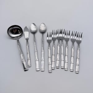 May include: A set of stainless steel flatware featuring a bamboo-inspired handle design. The collection includes one large soup ladle, one butter knife, two spoons, and six forks, all with a polished silver finish and segmented handle detailing.