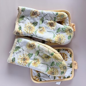 May include: A set of two oven mitts and a matching pot holder. The kitchen textiles feature a white background with a repeating pattern of yellow sunflowers and green leaves. The edges are trimmed with a yellow fabric.