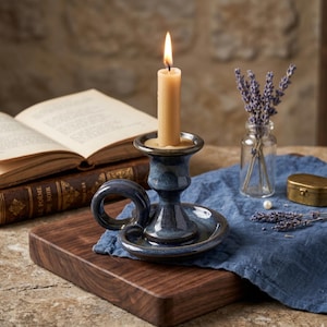May include: A lit candle sits in a blue ceramic candlestick holder with a handle. The holder is on a wooden tray with an open book, a small glass vase of lavender, and a gold box. A blue cloth is underneath.