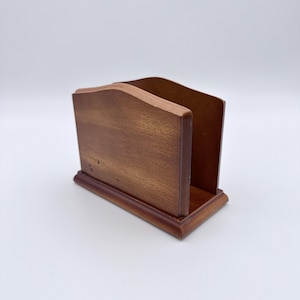 May include: A medium brown wooden napkin holder with a smooth finish and a curved top edge. The holder features a rectangular base with a beveled edge and two upright side panels connected by a central bottom piece to store napkins upright.