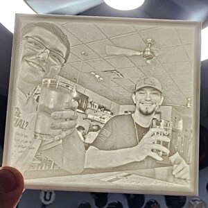 May include: A square, sepia-toned photo etched into a clear material, depicting two people holding glasses of a beverage. The background shows a bar setting with a ceiling fan and bar fixtures. The image is held by a hand.