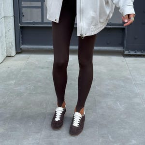 May include: A light-coloured jacket with a zip, worn over a light-coloured top and dark brown leggings. The outfit is paired with brown trainers with white laces. A brown handbag is held in the left hand.