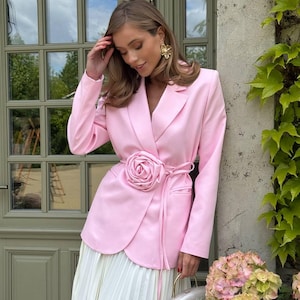 Peut inclure: Un blazer rose clair avec une ceinture en forme de rose, associé à une jupe plissée blanche. Le blazer a un col châle et des manches longues. Des boucles d'oreilles dorées et un sac à main argenté complètent la tenue. En arrière-plan, une porte verte et des plantes grimpantes.
