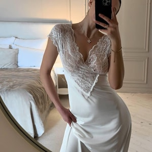 K&ouml;nnte beinhalten: Ein wei&szlig;es Slip-Kleid mit einem Spitzenoberteil und kurzen &Auml;rmeln. Das Kleid hat einen V-Ausschnitt und einen flie&szlig;enden Rock. Die Spitzendetails sind aufwendig und zart. Das Kleid ist aus einem weichen, seidigen Material.