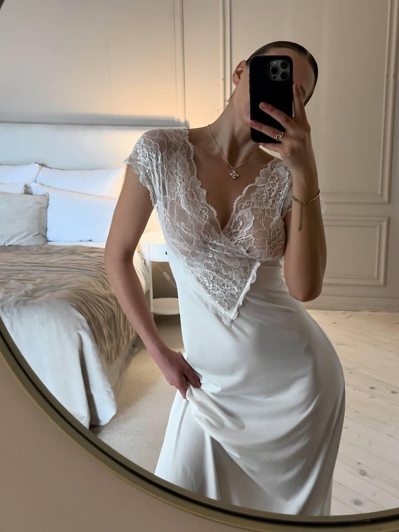 K&ouml;nnte beinhalten: Ein wei&szlig;es Slip-Kleid mit einem Spitzenoberteil und kurzen &Auml;rmeln. Das Kleid hat einen V-Ausschnitt und einen flie&szlig;enden Rock. Die Spitzendetails sind aufwendig und zart. Das Kleid ist aus einem weichen, seidigen Material.