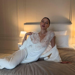K&ouml;nnte beinhalten: Ein wei&szlig;es Nachthemd mit Spitzendetails am Ausschnitt und an den &Auml;rmeln. Ein passender Morgenmantel ist &uuml;ber den Schultern drapiert. Die Dessous werden auf einem Bett mit einer neutralfarbenen Bettdecke und Kissen getragen.