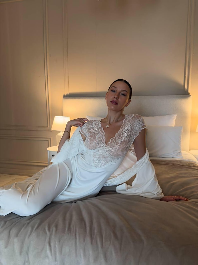 K&ouml;nnte beinhalten: Ein wei&szlig;es Nachthemd mit Spitzendetails am Ausschnitt und an den &Auml;rmeln. Ein passender Morgenmantel ist &uuml;ber den Schultern drapiert. Die Dessous werden auf einem Bett mit einer neutralfarbenen Bettdecke und Kissen getragen.