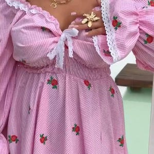 Puede incluir: Vestido a rayas rosas y blancas con bordado de rosas rojas. El vestido con hombros descubiertos presenta mangas largas y abullonadas con ribete de encaje blanco. Un lazo blanco adorna el escote. Collares dorados y un anillo de flores completan el look.