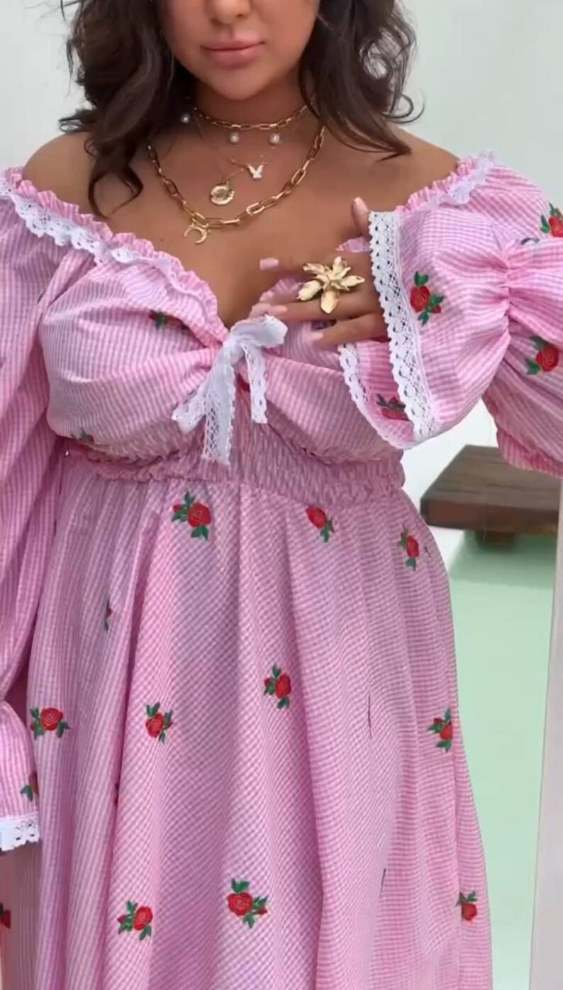 Puede incluir: Vestido a rayas rosas y blancas con bordado de rosas rojas. El vestido con hombros descubiertos presenta mangas largas y abullonadas con ribete de encaje blanco. Un lazo blanco adorna el escote. Collares dorados y un anillo de flores completan el look.
