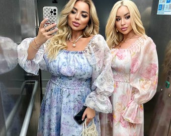 Puede incluir: Dos mujeres posan en un ascensor con vestidos estampados florales. Una lleva un vestido azul claro con estampado floral, la otra un vestido rosa floral. Ambos vestidos tienen mangas largas y transparentes y cinturas fruncidas.