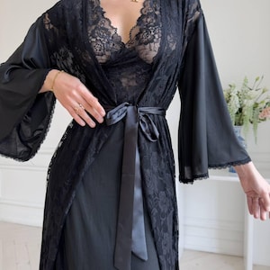 K&ouml;nnte beinhalten: Schwarzes Dessous-Set mit einer Spitzenrobe mit weiten &Auml;rmeln und einem Satinband. Das Set beinhaltet ein Spitzen-Bralette und ein Slip-Kleid, wodurch ein zusammenh&auml;ngendes und elegantes Ensemble entsteht. Die zarte Spitze verleiht dem Design einen Hauch von Raffinesse.