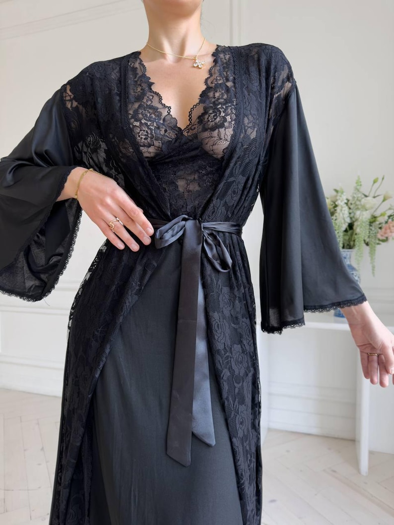 K&ouml;nnte beinhalten: Schwarzes Dessous-Set mit einer Spitzenrobe mit weiten &Auml;rmeln und einem Satinband. Das Set beinhaltet ein Spitzen-Bralette und ein Slip-Kleid, wodurch ein zusammenh&auml;ngendes und elegantes Ensemble entsteht. Die zarte Spitze verleiht dem Design einen Hauch von Raffinesse.