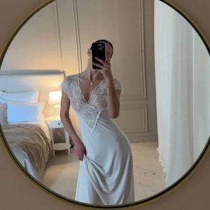 K&ouml;nnte beinhalten: Ein langes, elfenbeinfarbenes Slip-Kleid mit V-Ausschnitt und Spitzendetails am Oberteil. Das Kleid spiegelt sich in einem runden Spiegel mit goldenem Rahmen. Der Hintergrund umfasst ein Bett, einen Nachttisch und eine Wand.