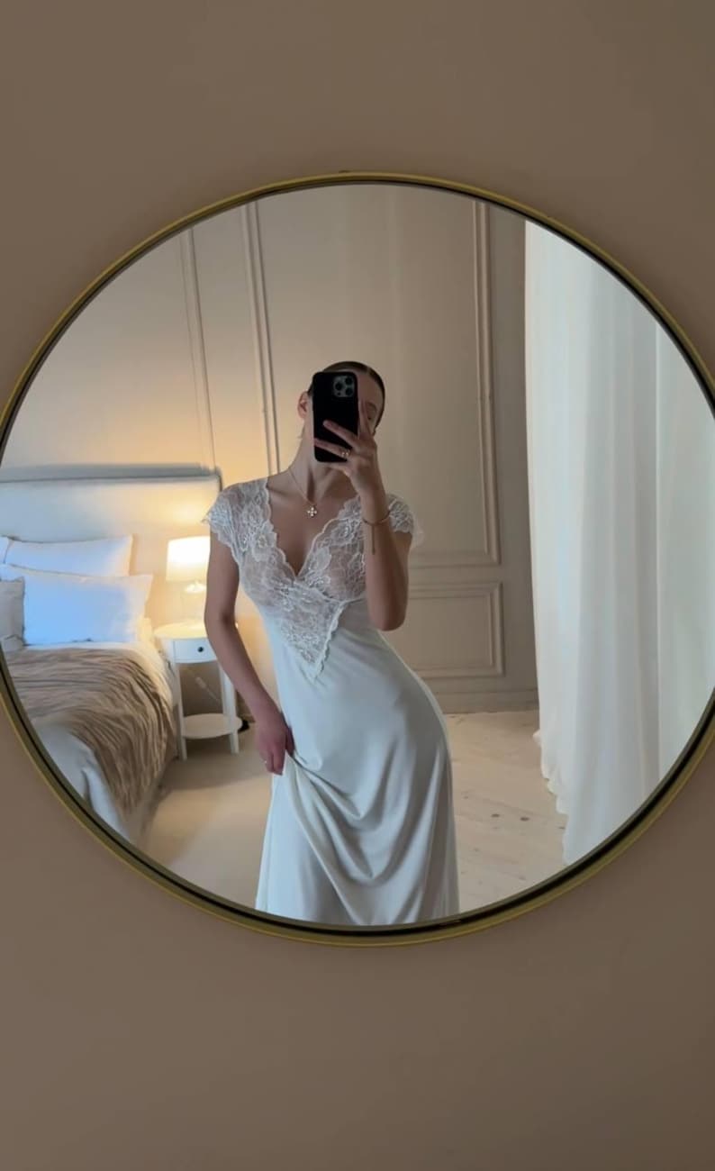 K&ouml;nnte beinhalten: Ein langes, elfenbeinfarbenes Slip-Kleid mit V-Ausschnitt und Spitzendetails am Oberteil. Das Kleid spiegelt sich in einem runden Spiegel mit goldenem Rahmen. Der Hintergrund umfasst ein Bett, einen Nachttisch und eine Wand.