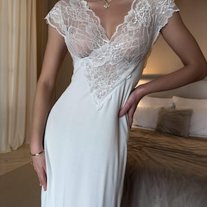 K&ouml;nnte beinhalten: Ein wei&szlig;es, bodenlanges Slip-Kleid mit V-Ausschnitt und kurzen &Auml;rmeln. Das Oberteil hat aufwendige wei&szlig;e Spitzendetails. Das Kleid ist aus einem weichen, flie&szlig;enden Stoff gefertigt. Eine goldene Halskette und ein Armband sind sichtbar.