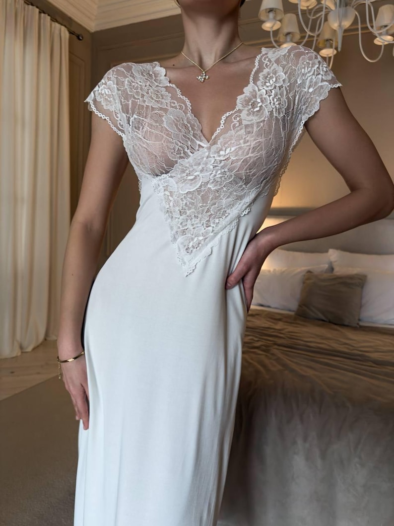 K&ouml;nnte beinhalten: Ein wei&szlig;es, bodenlanges Slip-Kleid mit V-Ausschnitt und kurzen &Auml;rmeln. Das Oberteil hat aufwendige wei&szlig;e Spitzendetails. Das Kleid ist aus einem weichen, flie&szlig;enden Stoff gefertigt. Eine goldene Halskette und ein Armband sind sichtbar.