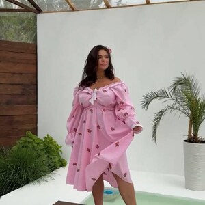 Puede incluir: Vestido rosa con hombros descubiertos, mangas largas y dobladillo a la rodilla. El vestido est&aacute; adornado con peque&ntilde;os estampados florales rojos y un lazo blanco en el escote. La modelo est&aacute; de pie sobre una plataforma de madera.