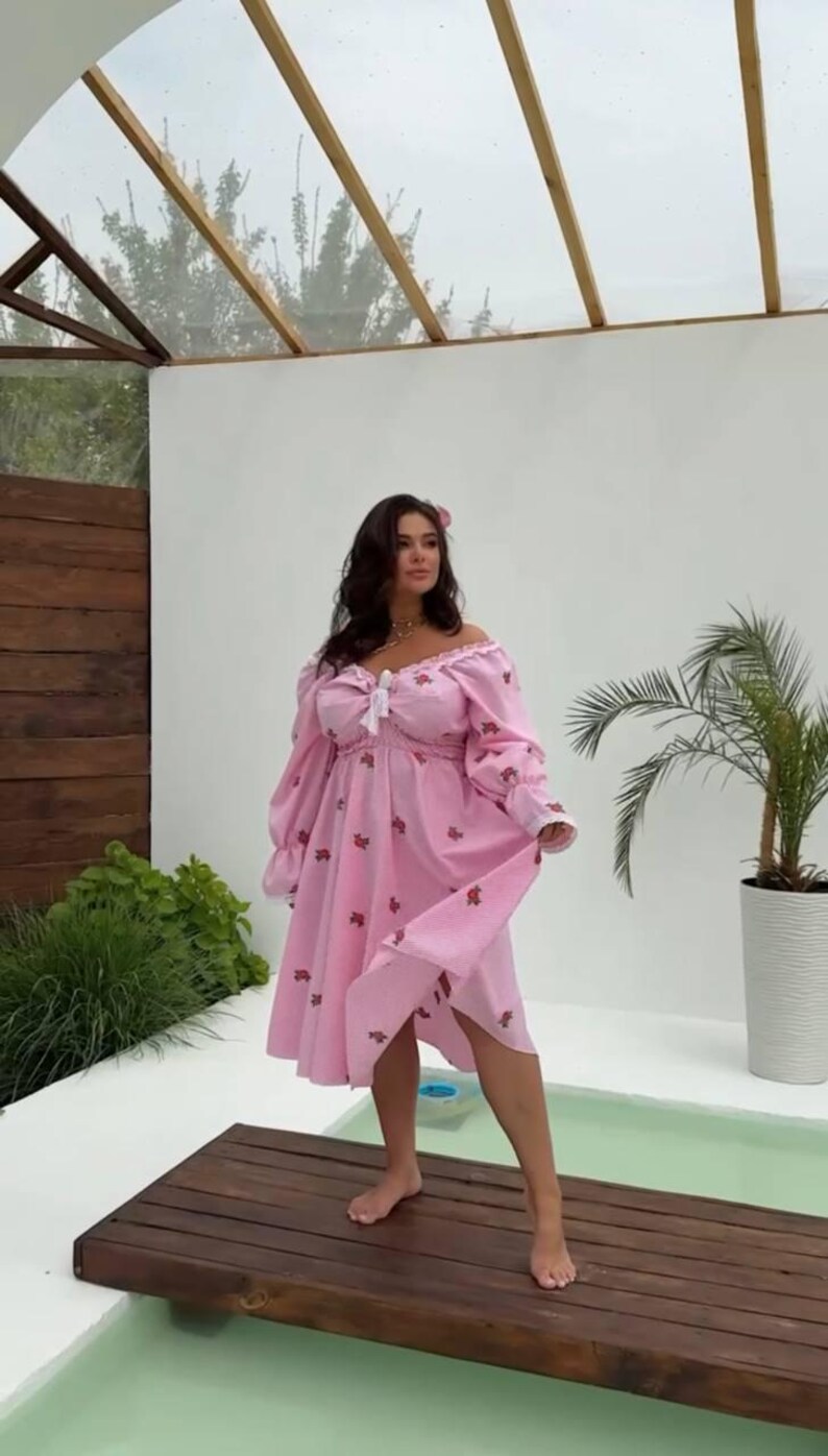 Puede incluir: Vestido rosa con hombros descubiertos, mangas largas y dobladillo a la rodilla. El vestido est&aacute; adornado con peque&ntilde;os estampados florales rojos y un lazo blanco en el escote. La modelo est&aacute; de pie sobre una plataforma de madera.