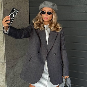 Könnte beinhalten: Ein dunkelgrauer Blazer mit doppelreihigem Design, kombiniert mit einem gestreiften Hemd und einer grauen Baskenmütze. Die Person trägt eine Sonnenbrille und hält ein Telefon. Das Outfit wird durch eine graue Handtasche ergänzt.