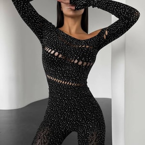 Könnte beinhalten: Schwarzer Jumpsuit mit langen Ärmeln und einem Cut-Out-Design. Der Jumpsuit ist mit kleinen, glitzernden Verzierungen bedeckt. Die Unterschenkel haben einen transparenten, gemusterten Stoff.