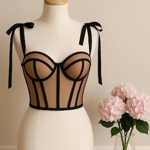 Puede incluir: Un corsé color canela con ribetes y tirantes negros, exhibido en un maniquí. El corsé presenta un diseño estructurado con líneas negras que delinean el busto y la cintura. Las correas de cinta negra se atan en los hombros. Un jarrón de flores rosas está al lado.