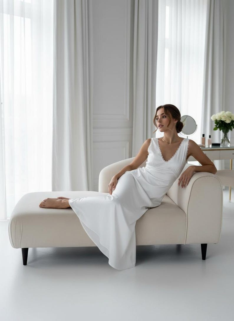 May include: A white, sleeveless dress with a V-neck and lace trim. The dress is worn by a person reclining on a cream-colored chaise lounge. The room has white curtains and a vanity with a mirror and flowers.