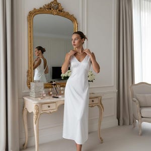 May include: A woman in a white slip dress stands in front of a gold-framed mirror. The dress has a V-neckline and lace trim. A vanity table with makeup and a bouquet of white flowers is visible.