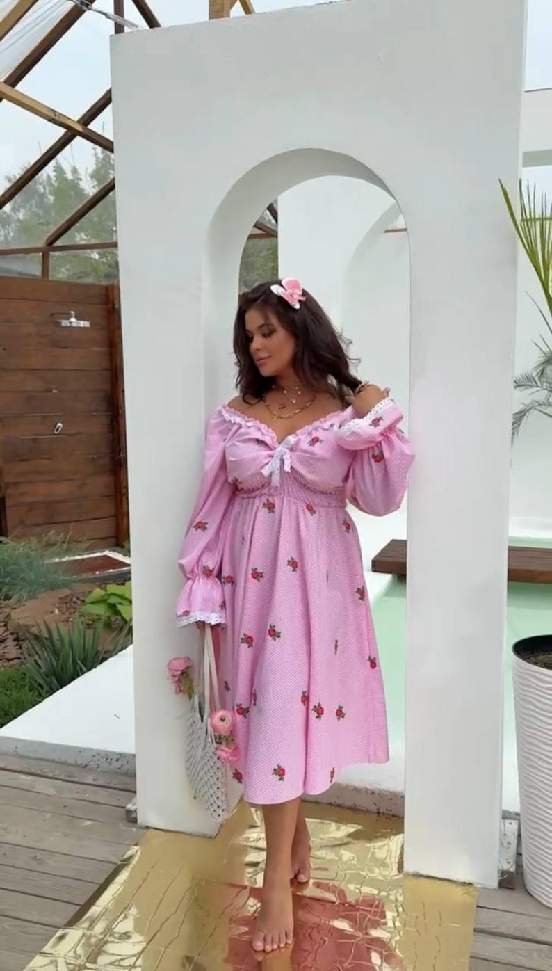Puede incluir: Vestido rosa con lunares blancos y detalles de rosas rojas. El vestido tiene mangas largas, escote coraz&oacute;n y un lazo. Se sostiene un bolso tote blanco con flores rosas.