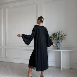 K&ouml;nnte beinhalten: Ein schwarzes Kleid mit weiten &Auml;rmeln und einem Spitzenbesatz. Das Kleid ist knielang und hat ein schlichtes, elegantes Design. Die &Auml;rmel sind lang und flie&szlig;end, und die Spitze sorgt f&uuml;r einen Hauch von Detail. Das Kleid wird mit transparenten Sandalen mit Absatz kombiniert.