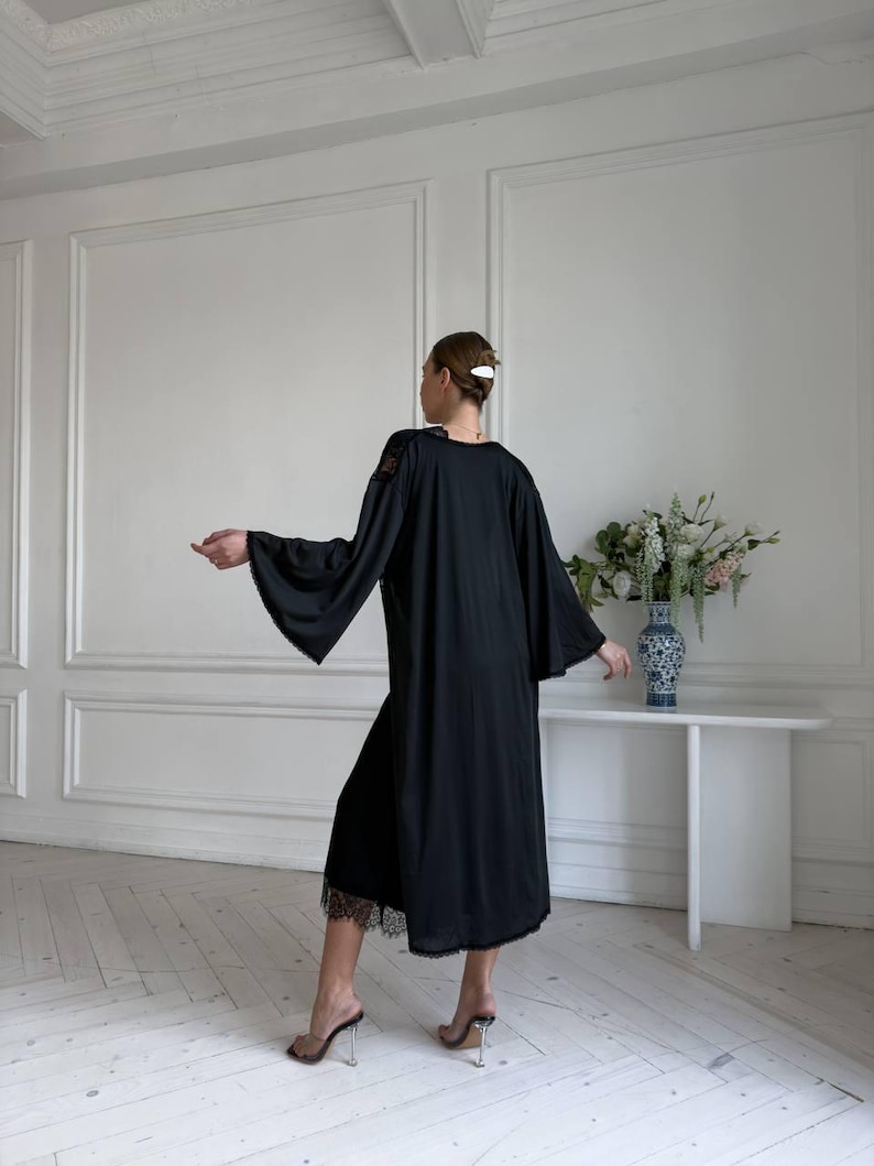 K&ouml;nnte beinhalten: Ein schwarzes Kleid mit weiten &Auml;rmeln und einem Spitzenbesatz. Das Kleid ist knielang und hat ein schlichtes, elegantes Design. Die &Auml;rmel sind lang und flie&szlig;end, und die Spitze sorgt f&uuml;r einen Hauch von Detail. Das Kleid wird mit transparenten Sandalen mit Absatz kombiniert.