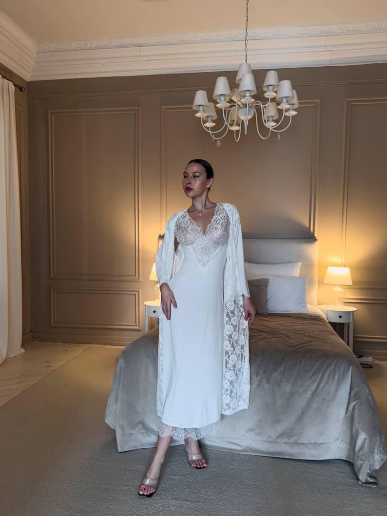 K&ouml;nnte beinhalten: Ein wei&szlig;es Satin-Nachthemd mit Spitzendetails am Oberteil und am Saum. Ein passender wei&szlig;er Bademantel mit Spitzen&auml;rmeln wird &uuml;ber dem Nachthemd getragen. Das Schlafzimmer hat eine neutrale Farbpalette mit einem Kronleuchter.