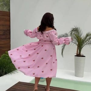 Puede incluir: Vestido rosa con hombros descubiertos y estampado floral. El vestido tiene mangas largas con pu&ntilde;os con volantes y cintura entallada. La modelo est&aacute; de pie sobre una plataforma de madera cerca de una piscina, con una palmera al fondo.
