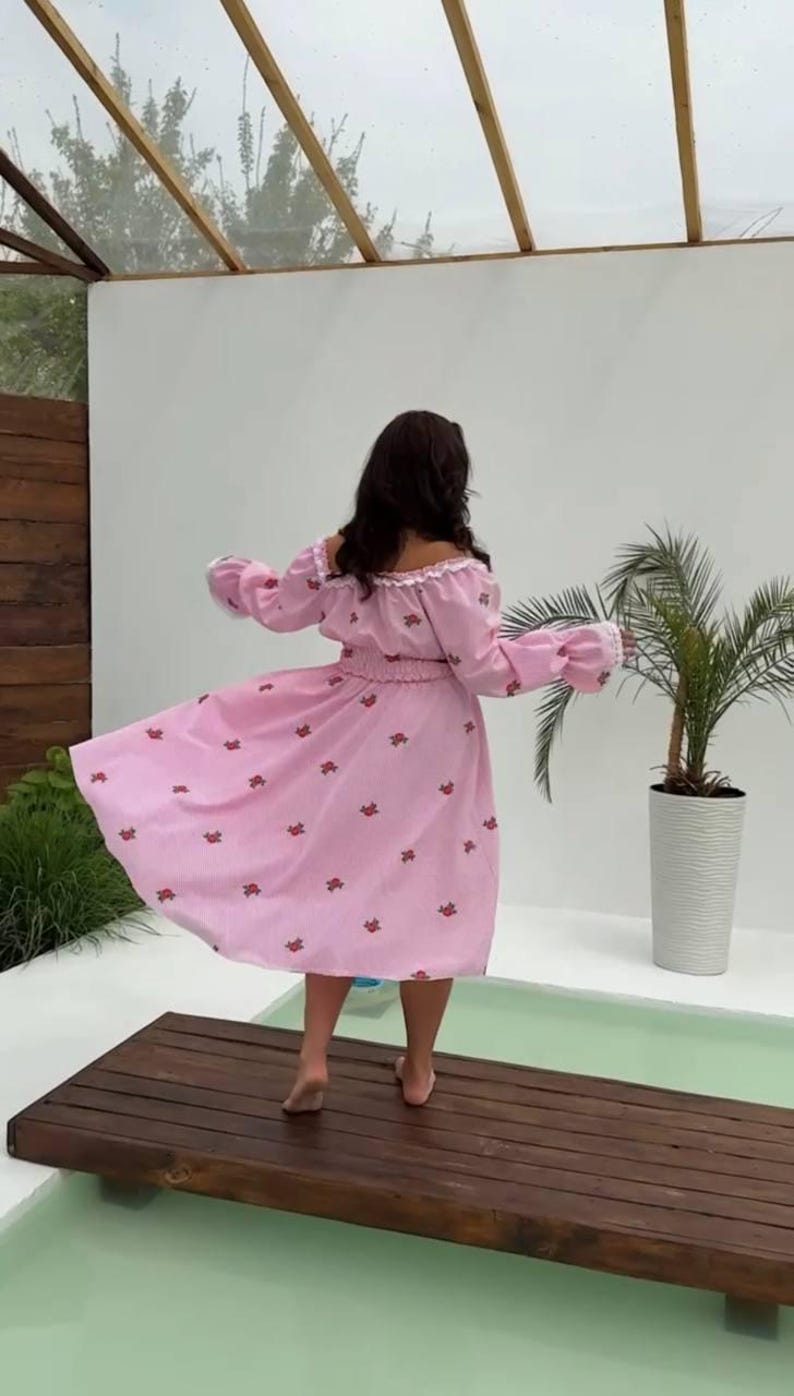 Puede incluir: Vestido rosa con hombros descubiertos y estampado floral. El vestido tiene mangas largas con pu&ntilde;os con volantes y cintura entallada. La modelo est&aacute; de pie sobre una plataforma de madera cerca de una piscina, con una palmera al fondo.